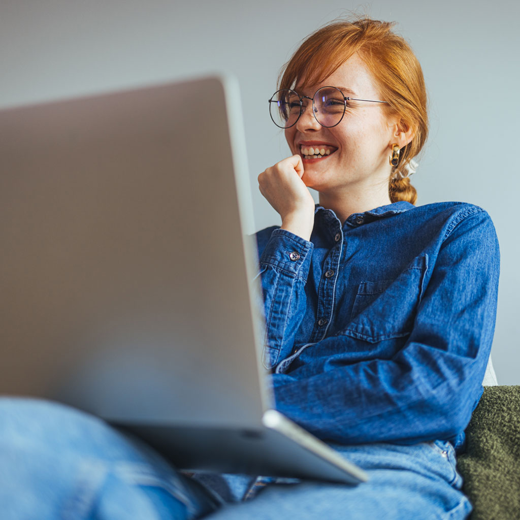 Erfreute junge Frau, die auf der Couch am Laptop sitzt.
